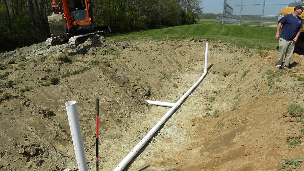 A Job site with a supervisor and a large dirt hole with pvc pipe vents being filled in with a backhoe